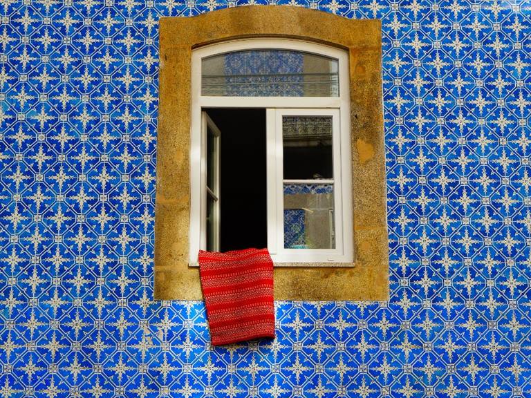 A Lisbon window set in weathered stone, framed by blue-and-white azulejo tiles, with a red cloth on the sill.