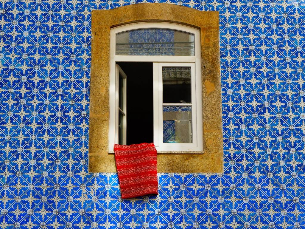 A Lisbon window set in weathered stone, framed by blue-and-white azulejo tiles, with a red cloth on the sill.