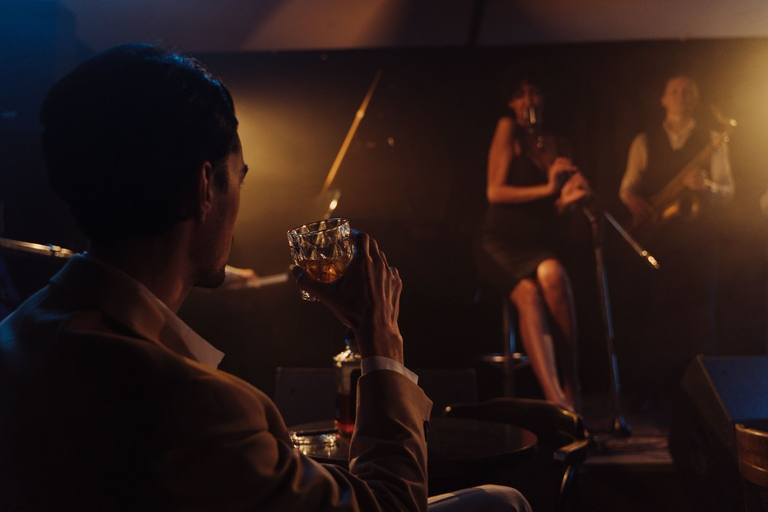 A listener holds a glass toward a small stage where a singer and bassist perform in warm club light.