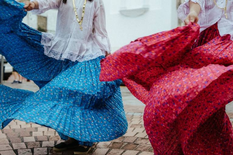 Two dancers in bright blue and pink skirts spinning on a sunlit street.