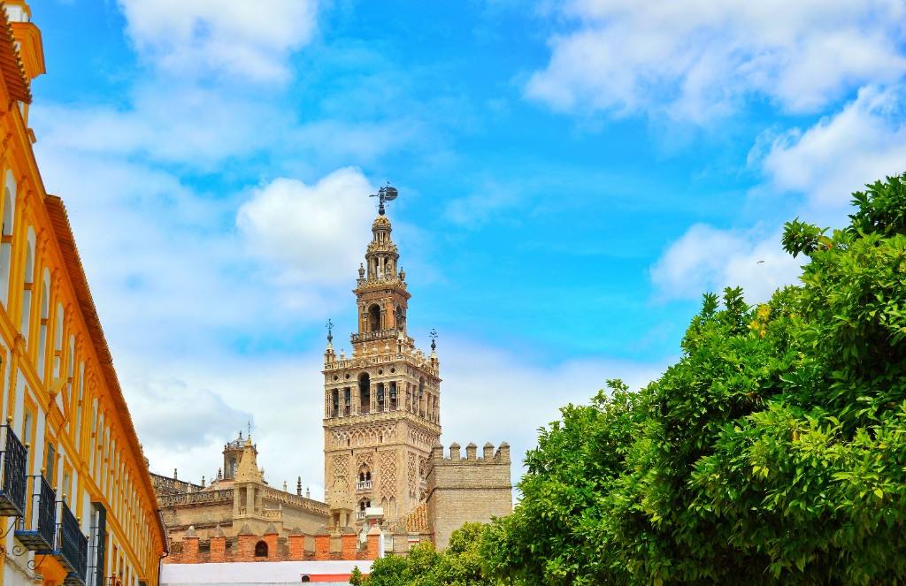 The Giralda rising above Seville Cathedral, framed by a yellow façade and green foliage under a blue sky.