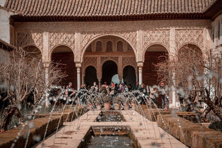 Uzun yansıtma havuzlu bir Granada avlusu; oymalı Nasrid kemerler ve cephe altında ziyaretçiler.