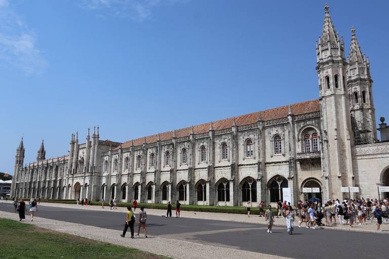 The long limestone façade of Jerónimos Monastery under a clear blue sky, with visitors along the paved front.