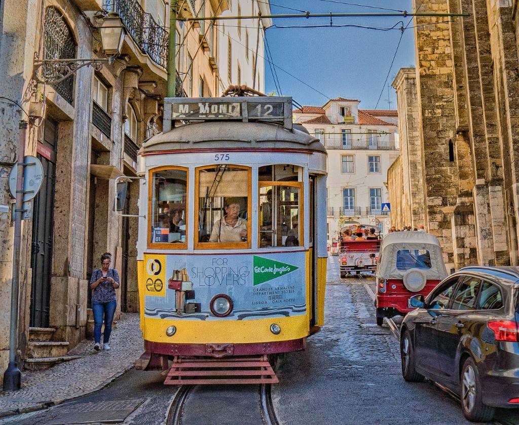 A yellow-and-white Lisbon tram descending a narrow cobbled street between stone façades under a clear sky.