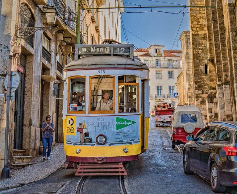 A yellow-and-white Lisbon tram descending a narrow cobbled street between stone façades under a clear sky.