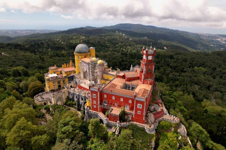 Pena Palace above the forested Sintra hills, with red and yellow Romantic towers under a cloudy sky.