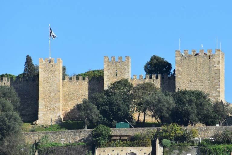 The stone towers and battlements of São Jorge Castle above green trees against a clear blue sky.