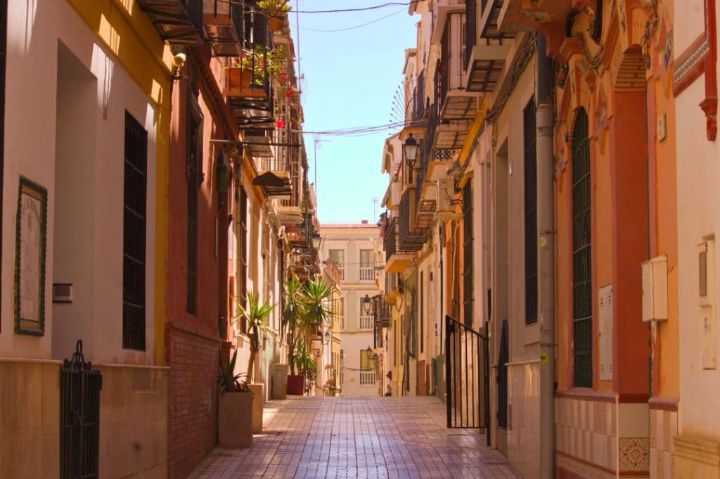 A narrow Seville alley in warm late light, with ochre walls, iron balconies and tiled pavements.