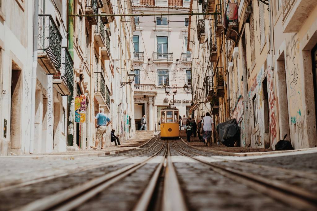 A yellow Lisbon tram climbing a steep cobbled street between cream façades and iron balconies.