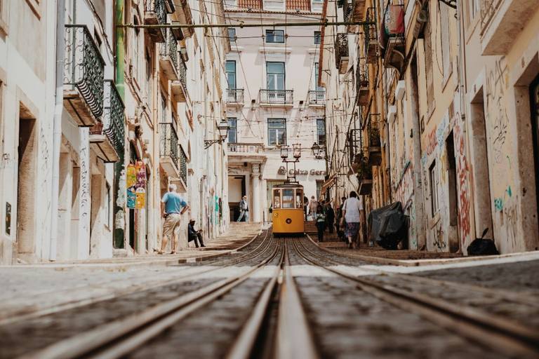 A yellow Lisbon tram climbing a steep cobbled street between cream façades and iron balconies.
