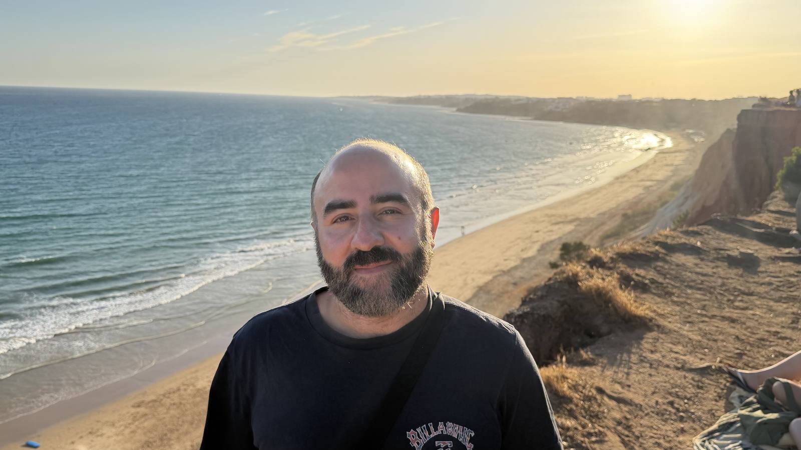 Yusuf standing on a coastal cliff above a long sandy beach.